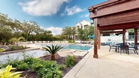 This expansive outdoor amenity space delivers a resort-style experience set against a backdrop of mature Texas live oak trees and natural Hill Country landscaping. Two generously sized swimming pools serve as the centerpiece of the grounds — one featuring a freeform design with natural limestone coping, decorative boulders, and tropical plantings including a striking sago palm, complemented by a full row of white lounge chairs along the pool deck. The second pool offers a classic rectangular layout bordered by a stone retaining wall and additional lounge seating, with a charming poolside structure featuring a red tile roof and an outdoor fireplace nearby. A bold wooden pergola provides shaded gathering space between the pools, outfitted with blue umbrellas and casual seating. Pathways of smooth concrete wind through native landscaping featuring wildflowers, ornamental grasses, and river rock accents. Limestone steps with modern steel railings lead to an elevated hammock area nestled among the trees, offering a tranquil retreat. A Spanish-style clubhouse structure with large glass windows and arched entryways anchors the space, providing indoor access to additional amenities. The overall atmosphere is relaxed, natural, and thoughtfully designed for outdoor living.