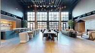 Located at 301 Winchester Ave in New Haven, CT, this expansive single-floor commercial space blends industrial architecture with refined modern design across a generously sized open floor plan. Soaring ceilings with exposed ductwork and dark structural elements contrast beautifully with warm wood slatted wall panels that appear throughout, lending the space a cohesive and polished aesthetic.

The main lounge area is anchored by dramatic floor-to-ceiling industrial-pane windows that flood the interior with abundant natural light and offer views of the surrounding neighborhood. Polished concrete floors run throughout, unifying the various zones. This central gathering area features varied seating arrangements, including modular sofas, curved lounge chairs, and a large communal table with pendant string lights overhead.

A stylish bar station, clad in slate-toned brick tile and topped with a granite-style surface, is framed by warm wood slat paneling with a backlit display niche — ideal for social gatherings. Nearby, a lounge nook with a linear fireplace set into the brick wall provides a welcoming, intimate atmosphere.

The entry reception is marked by a distinctive living moss wall behind a brick-faced desk. A long corridor lined with wood-slat-clad private rooms leads to a cozy private office, complete with built-in wood shelving and a round meeting table. Indoor trees placed throughout add organic warmth to the entire space.