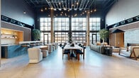 Located at 301 Winchester Ave in New Haven, CT, this expansive single-floor commercial space blends industrial architecture with refined modern design across a generously sized open floor plan. Soaring ceilings with exposed ductwork and dark structural elements contrast beautifully with warm wood slatted wall panels that appear throughout, lending the space a cohesive and polished aesthetic.

The main lounge area is anchored by dramatic floor-to-ceiling industrial-pane windows that flood the interior with abundant natural light and offer views of the surrounding neighborhood. Polished concrete floors run throughout, unifying the various zones. This central gathering area features varied seating arrangements, including modular sofas, curved lounge chairs, and a large communal table with pendant string lights overhead.

A stylish bar station, clad in slate-toned brick tile and topped with a granite-style surface, is framed by warm wood slat paneling with a backlit display niche — ideal for social gatherings. Nearby, a lounge nook with a linear fireplace set into the brick wall provides a welcoming, intimate atmosphere.

The entry reception is marked by a distinctive living moss wall behind a brick-faced desk. A long corridor lined with wood-slat-clad private rooms leads to a cozy private office, complete with built-in wood shelving and a round meeting table. Indoor trees placed throughout add organic warmth to the entire space.