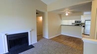 This single-floor residence offers a thoughtfully designed open-concept layout that combines comfort and functionality. The living area serves as the heart of the space, anchored by a classic white-mantled fireplace with a sleek black surround, adding a focal point of warmth and character. Vaulted ceilings rise overhead, enhancing the sense of openness and airiness throughout the room. Neutral-toned walls and light-colored trim create a clean, welcoming ambiance that complements the plush carpeting underfoot in the main living zone. Transitioning toward the kitchen area, the flooring shifts to smooth hardwood-style planks, providing a natural contrast. A ceiling fan with integrated lighting is centrally positioned to ensure comfort and illumination. The kitchen is partially open to the living space via a raised bar-height counter with a dark surface, offering a convenient pass-through and casual seating opportunity. White cabinetry and modern appliances, including a built-in microwave, are visible in the kitchen, contributing to a bright and efficient cooking space. A hallway connects to additional areas of the home, maintaining a cohesive and practical flow throughout the single-level floor plan.