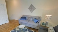 This two-floor space offers a well-appointed interior that blends modern style with homey comfort. The living area immediately sets the tone with warm hardwood flooring that extends throughout, providing a natural and inviting foundation for the space. A neutral-toned sofa serves as the centerpiece of the living area, accented with an array of blue and grey decorative pillows that add pops of color and texture. A geometric-patterned area rug in blue and white anchors the seating arrangement, complementing a glass-topped coffee table that adds a light, open feel to the room. Above the sofa, an ornate diamond-shaped wall medallion in grey serves as a striking focal point, bringing an artistic and decorative element to the otherwise clean, neutral walls. The layout is thoughtfully designed to maximize comfort and flow, with ample natural light contributing to the bright and airy ambiance. Across its two floors, the space provides a versatile layout suited to a variety of needs. The overall aesthetic is cohesive and contemporary, with carefully chosen furnishings and accents that create a warm, lived-in atmosphere that feels both functional and visually appealing.