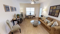 This single-floor space presents an open-concept living and dining area designed for both comfort and functionality. The living area is anchored by a glass-top coffee table with decorative ironwork base, complemented by a warm tan sofa adorned with patterned accent pillows. A floor lamp provides additional ambient lighting alongside the ceiling fan with integrated light fixture overhead, which serves both practical and aesthetic purposes.

The dining area sits prominently near a large window dressed with white plantation shutters, allowing natural light to filter softly into the space. A round bistro-style table with wrought-iron legs is surrounded by upholstered chairs, creating an intimate dining setting. A secondary seating arrangement near the entrance features dark wood furniture and accent chairs, adding versatility to the layout.

The space is finished with plush wall-to-wall carpeting in a warm beige tone, contributing to a cozy atmosphere throughout. Neutral cream-colored walls enhance the sense of openness, while framed artwork and decorative accents add character. A secondary door on the right side of the room offers additional access and brings in extra natural light, further brightening the interior.