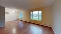 This single-floor unit in Essex offers a practical and straightforward layout with two bedrooms and one full bathroom. The living area is notably spacious, featuring warm hardwood flooring and large windows that invite natural light and provide outdoor views. A ceiling fan adds functionality to this generously sized room.

The galley-style kitchen is efficiently arranged with white cabinetry lining both walls, offering ample storage space. It includes a gas range, refrigerator, and a sink with counter space on the opposite wall. Light tile flooring and overhead lighting complete the kitchen's clean, utilitarian design.

Both bedrooms are carpeted and well-proportioned, each featuring sliding windows with outdoor views and baseboard heating. One bedroom includes a built-in closet with shelving, providing convenient storage.

The full bathroom features a tiled tub-and-shower combination with sliding glass doors, a mosaic tile floor, wainscoting-style tile walls, a toilet, and a vanity with sink. A frosted window allows natural light while maintaining privacy. Baseboard heating is present throughout the unit, ensuring consistent warmth across all rooms.