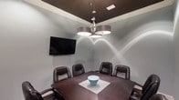 This single-level conference room offers a polished, professional setting designed for focused meetings and presentations. The space is anchored by a dark wood conference table surrounded by high-back executive leather chairs, providing a formal yet comfortable atmosphere for attendees. A wall-mounted flat-screen television serves as the primary display for presentations or video conferencing, positioned prominently for clear visibility from all seating positions. Overhead, a striking multi-drum pendant chandelier with a chrome finish provides the room's primary illumination, casting warm, directed light across the table while adding a refined design element to the space. The room features a two-tone color scheme, with soft light gray walls contrasted by a deep espresso-colored tray ceiling framed by crisp white crown molding. This ceiling detail adds architectural interest and a sense of depth. The overall ambiance is professional and intimate, making the space well-suited for small group meetings, client consultations, or collaborative work sessions.