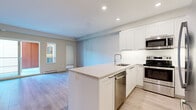 This well-designed two-bedroom, two-bathroom unit offers an efficient and functional layout across a single floor. The open-concept kitchen and living area forms the heart of the space, featuring light wood-style flooring that flows throughout. The kitchen is appointed with white shaker-style cabinetry extending to the ceiling, a stainless steel appliance package including a refrigerator, electric range, built-in microwave, and dishwasher, as well as a peninsula with a light stone-finish countertop that provides additional workspace and separates the kitchen from the living area. The living room receives natural light through a sliding glass door that opens onto a private balcony offering views of landscaped greenery and a forested hillside. Both bedrooms are finished with the same wood-style flooring and neutral walls, with the second bedroom featuring a sliding closet door. The two bathrooms are finished with white vanities, stone-surface countertops, undermount sinks, dark hardware accents, and glass-enclosed showers. Two hallways connect the rooms efficiently, and a dedicated closet provides additional storage within the unit.
