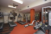 This fitness center is organized into distinct functional zones, creating a comprehensive workout environment across a single level. The space is divided into two primary areas: a cardio zone and a strength training zone, each thoughtfully designed to maximize usability.

The cardio area features an elongated layout lined with large windows along one wall, flooding the space with natural light and offering views of surrounding greenery. Equipment includes treadmills, stationary bikes, elliptical machines, and a cable machine positioned toward the far end. A bold, multi-colored striped carpet defines this zone, adding a dynamic visual element underfoot. Ceiling fans and overhead fluorescent lighting ensure comfort during workouts.

The strength training zone transitions to a solid orange carpet, creating a clear visual distinction between areas. A full dumbbell rack is positioned against a wall-length mirror, which enhances the sense of space. Additional strength equipment, a weight bench, and a barbell are also present. Ceiling-mounted flat-screen TVs are accessible from multiple areas throughout the facility.

Glass block accents, yellow and wood-toned wall details, and a glass door leading to an outdoor pathway contribute to the overall modern, energetic ambiance of the space.