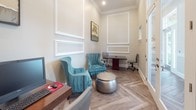 Located at 6450 Mercantile Dr E in Frederick, Maryland, this single-floor commercial office space combines professional functionality with refined interior design. The space is finished with distinctive herringbone-patterned hardwood-style flooring that runs throughout, lending a sophisticated character to every area. Walls are painted in a soft, neutral greige tone and adorned with crisp white decorative panel molding, adding architectural depth and elegance. Crown molding further enhances the polished aesthetic throughout the space. The main office area is thoughtfully arranged with a dark wood executive desk accompanied by ergonomic task chairs, creating a productive workstation. A seating area features plush teal velvet accent chairs with nailhead trim detail, paired with a metallic hammered-drum side table, offering a comfortable and stylish setting for clients or guests. Natural light filters through windows fitted with white blinds, brightening the interior and contributing to an inviting atmosphere. A glass-paneled interior door adds a sense of openness while maintaining defined spaces. The overall ambiance strikes a balance between professional efficiency and upscale design, making this office well-suited for client-facing business operations.