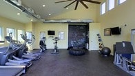 The fitness center at Lakewood Park Apartments is a well-equipped, single-floor facility designed to support a variety of workout styles. The space features soaring vaulted ceilings accented by a large ceiling fan, recessed lighting, and high windows that allow natural light to brighten the room, creating an open and energizing atmosphere.

The interior is finished with rubber flooring suited for both cardio and functional training activities. Cardio equipment, including stationary bikes and a stair climber, is arranged along one side of the room, while a wall-mounted flat-screen television provides entertainment during workouts. A prominent chalkboard wall serves as a focal point, offering space for workout tracking, class schedules, or motivational notes.

The functional training area includes a large tractor tire for strength and conditioning exercises, along with a rack of medicine and stability balls for versatile training options. An exposed ventilation duct runs across the ceiling, adding an industrial aesthetic to the space. The light yellow walls contribute to the room's bright, welcoming ambiance, making this fitness center a practical and dynamic amenity for residents.