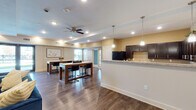 Located at 4702 Ambassador Way in Balch Springs, Texas, this single-story community space offers an open-concept floor plan designed for comfort and versatility. Rich hardwood-style flooring flows throughout, anchoring each area with warmth and visual continuity.

The kitchen is a standout feature, outfitted with dark espresso cabinetry, light granite countertops, and ample workspace arranged in a U-shaped configuration. Pendant lighting adds a refined touch, while generous counter and cabinet space make it well-suited for both everyday use and larger gatherings. A breakfast bar peninsula connects the kitchen to the adjacent dining area, where a high-top table with bar seating sits beneath a ceiling fan with recessed lighting above.

The lounge area serves as the heart of the space, centered around a classic fireplace framed by a crisp white mantle and flanked by built-in shelving units that offer both display and storage space. The room is well-lit with recessed lighting and accommodates multiple seating arrangements, making it ideal for social gatherings or relaxed conversation.

Natural light filters in through windows near the entry, and the layout transitions seamlessly between zones. Warm neutral wall tones, consistent flooring, and carefully considered lighting create a cohesive, welcoming ambiance throughout this well-appointed community space.