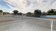 This outdoor tennis facility offers multiple full-sized hard courts, each equipped with regulation nets and clearly marked court lines. The courts feature a two-tone surface with a lighter playing area and darker red-clay-colored service boxes, providing clear visual contrast for players. Perimeter chain-link fencing encloses the entire facility, with dark windscreen panels lining the outer boundaries to reduce wind interference and provide a defined backdrop for play. Seating benches are positioned courtside, offering convenient rest areas between games. Tall light poles are installed throughout the complex, indicating the courts are suitable for evening play. A covered pavilion with a distinctive hipped roof is situated adjacent to the courts, providing a shaded gathering space for spectators and players alike. The facility is surrounded by lush, mature tropical trees — including palms and broad-canopy hardwoods — that frame the courts and contribute to a pleasant, park-like setting. The open-air design allows for natural ventilation and unobstructed views of the sky, creating an inviting environment for recreational and competitive tennis. A paved parking area is accessible nearby, adding to the convenience of the facility.