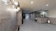 Located at 1539 Beloit Ave in the Sawtelle neighborhood of Los Angeles, this building's lobby presents a polished, contemporary aesthetic designed to make a strong first impression. The entrance corridor is lined with textured stone-look wall panels in warm gray tones, lending a sophisticated, upscale character to the space. Recessed lighting illuminates the area evenly throughout, complementing a sculptural cluster of globe pendant lights near the entrance that serve as a striking focal point.

The reception area is thoughtfully designed, featuring a low-profile desk wrapped in matching stone-patterned cladding and enclosed by sleek glass panels, creating a semi-private, professional atmosphere. Decorative white ceramic vases and floral accents add subtle elegance to the desk area.

A dedicated elevator lobby houses two stainless steel elevators set against the same stone-paneled walls, providing convenient vertical access throughout the building. The flooring transitions between plush neutral carpet and polished dark tile, defining distinct zones within the lobby. At the far end of the corridor, a glass door opens to an outdoor area, allowing natural light to filter in and enhancing the sense of depth and openness. Overall, the lobby exudes a clean, modern sensibility with carefully curated finishes and a calm, welcoming ambiance.