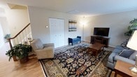 This two-floor home offers a thoughtfully arranged layout that balances comfort and functionality. The main living area sets the tone for the entire space, showcasing warm hardwood flooring that extends throughout and neutral, light-toned walls that create a bright and inviting atmosphere. A large, ornate area rug with rich navy, gold, and earth-toned patterns anchors the living room, adding a touch of elegance and warmth underfoot. The room is generously sized, providing ample space for seating and relaxation, with a plush sofa and a classic wooden coffee table complementing the traditional aesthetic. A large flat-screen television sits atop a sturdy wooden media console, making the living room well-suited for both entertainment and leisure. A closet with classic paneled doors offers convenient built-in storage. A stylish floor lamp provides additional ambient lighting, while wall art adds a decorative focal point. The space flows naturally across two floors, suggesting additional living, sleeping, or functional areas above or below. Throughout, the design leans toward a clean, traditional style with practical furnishings and a neutral palette that makes the home feel welcoming and move-in ready.