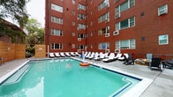This outdoor amenity space at 516 W Ormsby Ave in Louisville, Kentucky, is a well-appointed pool and recreation area nestled within the courtyard of a classic multi-story red brick building. The pool itself is generously sized with clear, light blue water and is surrounded by a smooth concrete deck that provides ample room for relaxation and movement. Numerous cushioned lounge chairs are arranged throughout the deck, offering comfortable spots for sunbathing and leisure.

A natural cedar wood fence encloses one side of the courtyard, providing privacy and a warm aesthetic contrast to the surrounding brick facade. Mounted along this cedar wall is a stainless steel grill, along with safety equipment including a rescue board and hose, ensuring both convenience and preparedness for pool users. Posted pool rules and signage are clearly visible along this wall.

Toward one end of the deck, a seating arrangement with Adirondack-style chairs surrounds a fire pit, creating an inviting social gathering spot. The enclosure formed by the multi-story brick building walls gives the space a sheltered, semi-private feel, while mature trees visible beyond the fence add a natural touch. The overall ambiance is that of a resort-style retreat set within an urban Louisville neighborhood.