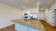 Located at 1909 Boynton Way in Bolivia, NC, this well-appointed single-story home offers three bedrooms, two bathrooms, and an attached garage across a comfortable and efficiently designed floor plan. The open-concept kitchen and living area serves as the heart of the home, showcasing a large granite-topped island with an undermount sink, white shaker-style cabinetry, a tile backsplash, and a full suite of stainless steel appliances including a range, over-the-range microwave, and French door refrigerator. Wood-look plank flooring flows throughout the main living areas, adding warmth and continuity. A central hallway connects the bedrooms and bathrooms, maintaining a clean and organized layout. The bedrooms are carpeted for comfort, with natural light streaming through large windows. Each bedroom includes closet space with wire shelving, and the primary bedroom is generously sized. The bathrooms feature wood-look flooring, white vanity cabinetry, and a tub-shower combination. The attached garage provides convenient interior access and includes an automatic opener. Recessed lighting throughout the home and a bright, neutral color palette create a clean, modern ambiance suited to a variety of lifestyles.