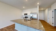 Located at 1909 Boynton Way in Bolivia, NC, this well-appointed single-story home offers three bedrooms, two bathrooms, and an attached garage across a comfortable and efficiently designed floor plan. The open-concept kitchen and living area serves as the heart of the home, showcasing a large granite-topped island with an undermount sink, white shaker-style cabinetry, a tile backsplash, and a full suite of stainless steel appliances including a range, over-the-range microwave, and French door refrigerator. Wood-look plank flooring flows throughout the main living areas, adding warmth and continuity. A central hallway connects the bedrooms and bathrooms, maintaining a clean and organized layout. The bedrooms are carpeted for comfort, with natural light streaming through large windows. Each bedroom includes closet space with wire shelving, and the primary bedroom is generously sized. The bathrooms feature wood-look flooring, white vanity cabinetry, and a tub-shower combination. The attached garage provides convenient interior access and includes an automatic opener. Recessed lighting throughout the home and a bright, neutral color palette create a clean, modern ambiance suited to a variety of lifestyles.
