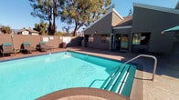 This outdoor amenity area is thoughtfully designed to provide a comprehensive recreational experience. At its center lies a generously sized swimming pool with clear turquoise water, complemented by a separate spa for relaxation. The pool deck is finished in stamped concrete, providing a durable and visually appealing surface that surrounds both the pool and spa.

A dedicated lounge zone features a stone fire pit encircled by Adirondack chairs in white and yellow, creating a welcoming gathering spot. Nearby, a pergola structure with teal curtain panels offers a shaded retreat, furnished with additional seating and tables beneath green umbrellas. Rows of chaise lounges are positioned along the pool's perimeter, ideal for sunbathing.

An adjacent single-story clubhouse building with a pitched roofline and stucco exterior connects seamlessly to the outdoor space via large glass sliding doors and floor-to-ceiling windows, allowing natural light to flow freely and providing direct views of the pool area. A covered patio with outdoor dining furniture extends from the building, bridging the indoor and outdoor environments.

The entire space is framed by mature trees, including tall pines and palms, with distant mountain views visible on the horizon under open blue skies, contributing to a serene, resort-like atmosphere.