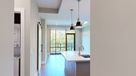This thoughtfully designed one-bedroom, two-bathroom residence at 350 Carraway Crossing in Chapel Hill, NC offers a refined living experience through modern architecture and high-quality finishes throughout. The open-concept kitchen and living area forms the heart of the home, anchored by a large center island with a white quartz countertop, undermount sink, and stylish pendant lighting. Cabinetry combines white upper shaker-style cabinets with blue-gray lower units, complemented by stainless steel appliances and glass-front accent cabinets. Light hardwood flooring runs throughout the main living area, and a sliding barn door adds a contemporary design touch. Floor-to-ceiling sliding glass doors in the living area flood the space with natural light and open to a balcony with wooded views. The spacious bedroom features plush carpeting, a ceiling fan, and its own large window framing lush tree canopies. The primary bathroom is a standout, showcasing a walk-in shower with floor-to-ceiling marble-style tile, a built-in bench, and frameless glass enclosure. A second bathroom offers white cabinetry with dark hardware and stone countertops. A dedicated closet and hallway complete the well-organized layout of this elegant Chapel Hill residence.