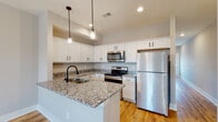 This two-story space is thoughtfully designed with contemporary finishes and a clean, neutral palette throughout. The kitchen serves as a focal point on the main level, showcasing white shaker-style cabinetry with matte black hardware, granite countertops with subtle veining, and a peninsula with an undermount stainless steel sink fitted with a black gooseneck faucet. Stainless steel appliances, including a refrigerator, electric range, and over-the-range microwave, complement the space, while pendant lights with glass globe fixtures add a decorative touch overhead. Warm-toned hardwood flooring runs throughout the main level, extending into a hallway lined with recessed lighting and paneled white doors.

The upper level transitions to plush carpeted bedrooms, each offering a calm, neutral atmosphere with ceiling fans and paneled doors leading to closets and connecting spaces. The bathrooms are finished with wood-look flooring, white vanities with stone-topped counters, and clean white surround showers — one featuring a standalone shower stall with built-in shelving, another offering a tub-shower combination. Throughout, matte black hardware provides a consistent, modern design accent.