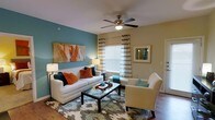 This single-floor residence offers a thoughtfully designed living space that balances color and comfort. The living room serves as the focal point of the home, showcasing a striking teal accent wall paired with warm, neutral-toned walls that create a lively yet inviting atmosphere. A ceiling fan with a light fixture anchors the room from above, providing both airflow and ambient lighting. Hardwood-style flooring runs throughout, adding warmth and a sense of continuity to the space. Natural light filters in through a large window dressed with striped curtains in neutral and gold tones, brightening the room and highlighting the colorful furnishings below. The seating arrangement features a white sofa adorned with vibrant teal and burnt orange accent pillows, complemented by a cream armchair and a sleek glass-topped coffee table resting on a multi-toned area rug. A bold abstract canvas painting on the accent wall adds an artistic touch, while a framed artwork on the adjacent wall contributes to the curated feel of the room. The overall ambiance is modern and welcoming, with a carefully chosen color palette of teal, orange, and gold that gives the space a dynamic yet cohesive character.