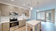This thoughtfully designed one-bedroom, one-bathroom residence offers a cohesive modern aesthetic throughout its efficiently utilized single-floor layout. Light-toned hardwood flooring runs continuously across the main living areas and bedroom, creating a warm and unified feel.

The open kitchen is a standout feature, showcasing flat-front cabinetry in a warm wood finish, white subway tile backsplash, and a full suite of stainless steel appliances including a refrigerator, range, microwave, and dishwasher. Pendant lighting hangs above a central white kitchen island, adding both style and functional prep space. The kitchen flows naturally into the living area, which benefits from a large window that invites ample natural light.

The bedroom is a generously sized retreat finished in soft neutral tones with light-washed walls, hardwood flooring, and a ceiling fan with light fixture. A large window with blinds provides natural light and ventilation, while a dedicated closet offers convenient storage.

The bathroom is clean and contemporary, featuring a wood-toned vanity with a white quartz countertop, a large mirror, and a glass-enclosed walk-in shower with white tile surround. Light tile flooring and recessed lighting complete the polished, spa-like atmosphere. A separate closet adjacent to the bathroom adds additional organizational space to the unit.