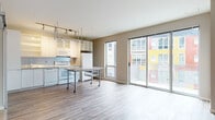 This 1-bedroom, 1-bathroom unit is designed with a clean, modern aesthetic and an open-concept floor plan that seamlessly connects the kitchen and living areas. The kitchen is well-appointed with white cabinetry featuring glass-front upper doors, granite countertops, white appliances including a range, dishwasher, microwave, and refrigerator, and a dedicated overhead track lighting system paired with an industrial-style wire rack for added storage. A movable kitchen island with a granite surface provides flexible workspace and dining options. Large sliding glass doors flood the living area with natural light and open to a private balcony overlooking the vibrant, colorful exterior of the surrounding building. Rich hardwood-style flooring runs throughout the main living spaces, adding warmth and continuity. The bedroom is generously proportioned, featuring exposed utility piping as an industrial design accent, a sliding barn-style door, and access to closet space. The bathroom offers a full tub-shower combo with white tile surround, a granite-topped vanity, geometric-patterned tile flooring, and built-in open shelving for convenient storage. This unit combines contemporary finishes with functional design for comfortable, modern living.