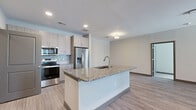 Located at 9445 FM-1464 N in Houston, TX, this single-floor, two-bedroom, two-bathroom unit offers a well-designed layout with contemporary finishes throughout. The open-concept kitchen and living area forms the heart of the space, featuring light wood-style luxury vinyl plank flooring, recessed lighting, and a ceiling fan. The kitchen is equipped with stainless steel appliances—including a refrigerator with ice maker, range, and microwave—complemented by light wood cabinetry that extends to the ceiling for ample storage. A central island with a granite countertop and undermount sink provides additional workspace and separates the kitchen from the living area. The living room receives natural light through windows and a glass door that opens to the exterior. Both bathrooms are finished with granite-topped vanities, wood-tone cabinetry, and tile flooring. One bathroom features a soaking tub with full marble-style tile surround. The bedrooms are carpeted and equipped with ceiling fans; each includes access to a walk-in closet with wire shelving systems. The primary bedroom is generously proportioned and benefits from natural light. The overall aesthetic is clean, neutral, and modern throughout.