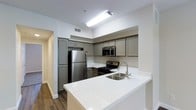 This single-floor residence offers a thoughtfully designed interior that balances modern aesthetics with functional living. The kitchen stands out as a central highlight, showcasing gray shaker-style cabinetry that extends to the upper walls, providing generous storage throughout. Stainless steel appliances — including a full-size refrigerator, gas range, and built-in microwave — lend a sleek, contemporary feel to the space. A crisp white quartz peninsula countertop extends outward, creating an ideal surface for casual dining or meal preparation, while a stainless steel undermount sink with a modern arc faucet adds a refined finishing touch. The warm-toned hardwood flooring grounds the space and contrasts elegantly with the bright white walls and countertops, creating a clean and inviting atmosphere. Recessed ventilation and flush ceiling lighting keep the kitchen bright and well-ventilated. A hallway visible from the kitchen suggests a well-connected floor plan with additional living spaces beyond. Overall, the residence presents a cohesive, move-in-ready environment that combines practical design with contemporary style, making it a comfortable and visually appealing place to call home.