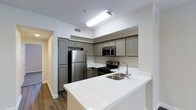 This single-floor residence offers a thoughtfully designed interior that balances modern aesthetics with functional living. The kitchen stands out as a central highlight, showcasing gray shaker-style cabinetry that extends to the upper walls, providing generous storage throughout. Stainless steel appliances — including a full-size refrigerator, gas range, and built-in microwave — lend a sleek, contemporary feel to the space. A crisp white quartz peninsula countertop extends outward, creating an ideal surface for casual dining or meal preparation, while a stainless steel undermount sink with a modern arc faucet adds a refined finishing touch. The warm-toned hardwood flooring grounds the space and contrasts elegantly with the bright white walls and countertops, creating a clean and inviting atmosphere. Recessed ventilation and flush ceiling lighting keep the kitchen bright and well-ventilated. A hallway visible from the kitchen suggests a well-connected floor plan with additional living spaces beyond. Overall, the residence presents a cohesive, move-in-ready environment that combines practical design with contemporary style, making it a comfortable and visually appealing place to call home.
