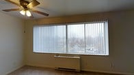 This single-floor space is defined by a standout architectural feature: a wide, expansive picture window fitted with vertical blinds that span nearly the full width of the wall. The window allows an abundance of natural light to fill the room, creating a bright and airy atmosphere throughout the day. Views through the glass extend outward to surrounding greenery and a street-level scene below, providing a pleasant visual connection to the outside environment.

The interior maintains a clean, neutral aesthetic with light-toned walls and flooring that contribute to an open, uncluttered feel. A ceiling fan is installed overhead, offering both air circulation and supplemental lighting, making the space practical across seasons. Baseboard heating runs beneath the large window, ensuring efficient climate control during colder months.

The overall ambiance of the space is functional and understated, with its most notable quality being the generous window that serves as a focal point. The combination of natural light, practical heating and cooling systems, and simple finishes makes this a versatile space well-suited for a variety of uses.