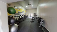 This fitness center in Mill Creek, Washington, is a single-floor facility designed to support a comprehensive workout experience. The space is divided into distinct functional zones, each thoughtfully appointed with appropriate equipment and finishes.

The main exercise area features durable rubber flooring and is lined with an impressive array of cardio machines, including multiple ellipticals and treadmills. A multi-station cable and weight machine anchors the strength training section, complemented by a full dumbbell rack offering a wide range of free weights and stability balls for functional training. Bold motivational typography covers the walls in black and yellow, with phrases encouraging perseverance and intensity, creating an energizing atmosphere throughout.

A dedicated spin and yoga studio offers a quieter, more versatile workout environment. This room features rich, dark hardwood flooring, neutral gray walls, and large windows with blinds that allow for natural light. A wall-mounted flat-screen television and ceiling fan add to the comfort, while French doors provide convenient access to the outdoors.

Recessed lighting and ceiling-mounted fixtures illuminate both areas effectively. The overall design is clean, modern, and focused, providing residents with a motivating environment to meet a wide range of fitness goals.