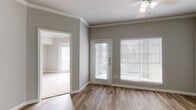 This single-floor residence offers a thoughtfully designed layout that balances comfort and functionality. The open living area is anchored by warm wood-look flooring that extends throughout the main spaces, complemented by neutral gray walls and crisp white crown molding that add a refined architectural touch. A ceiling fan provides additional comfort to the airy living space, which benefits from generous natural light streaming through large windows fitted with white blinds. A secondary room off the living area features access to the outdoors through a glass-paneled door, further enhancing the sense of openness.

The kitchen is a standout feature, showcasing bright white shaker-style cabinetry with brushed nickel hardware, stone-look countertops, and black appliances including an electric range, over-range microwave, and refrigerator. A peninsula counter separates the kitchen from the main living area, creating a convenient and social layout.

The bathroom is well-appointed with a dual-sink vanity topped with matching stone-look counters, white cabinetry, a wide illuminated mirror, and a full tub-shower combination. Wood-look flooring continues here, tying the space together cohesively. Throughout, the home maintains a clean, modern aesthetic with a neutral palette that offers a versatile and welcoming atmosphere.