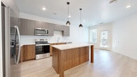 Located at 13500 Antioch Rd in Overland Park, KS, this thoughtfully designed one-bedroom, one-bathroom residence offers a clean, modern aesthetic across a single well-organized floor. The open-concept living and kitchen area serves as the heart of the home, showcasing light hardwood-style flooring, crisp white walls, and abundant recessed lighting. The kitchen is appointed with a full suite of stainless steel appliances — including a refrigerator, range, microwave, and dishwasher — complemented by two-tone cabinetry featuring gray upper cabinets and warm walnut-toned lower cabinets, a white quartz countertop, and a classic subway tile backsplash. A substantial center island with matching walnut paneling provides additional prep space and is illuminated by a pair of modern pendant lights. Natural light filters in through a glass door that opens toward an exterior area. The generously sized primary bedroom features plush carpeting, a ceiling fan with light, and a window that fills the room with daylight. The bathroom is finished with a wood-toned vanity, white quartz countertop, a large mirror, and a glass-enclosed shower, with direct access to a walk-in closet outfitted with wire shelving. A dedicated laundry area completes the functional layout of this well-appointed residence.
