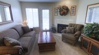 This single-floor residence offers a comfortable and well-appointed living space designed with warmth and functionality in mind. The living room serves as the heart of the home, anchored by a classic white-mantled fireplace that provides a focal point and a cozy atmosphere. Soft, neutral-toned carpeting runs underfoot, complementing the muted green walls and creating an overall sense of calm. A glass-paned exterior door, flanked by a large window fitted with blinds, allows generous natural light to pour into the room while offering convenient access to the outdoors. The space is thoughtfully arranged with comfortable seating, including a plaid sofa and an upholstered armchair, positioned around a rich wood coffee table with decorative ironwork detailing. A lush houseplant adds a touch of greenery near the door, enhancing the natural ambiance. Framed artwork and a decorative wreath adorn the walls above the fireplace, adding character and a personal touch. The overall layout is open yet intimate, making this living area well-suited for both relaxed everyday living and casual entertaining.
