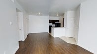 This single-floor residence offers an open-concept layout that seamlessly connects the living area to the kitchen, creating a spacious and functional living environment. The floors throughout the main living space are finished with rich, dark hardwood-style planks that contrast cleanly against the bright white walls, lending the interior a modern and polished feel.

The kitchen is positioned centrally within the open floor plan and features dark espresso-toned cabinetry, black appliances, and a white peninsula that serves as a natural divider between the cooking and living zones. The peninsula also provides additional counter space and a casual dining or prep area. The kitchen flooring transitions to a lighter tile or vinyl surface, clearly delineating the cooking space from the main living area.

Recessed lighting is installed throughout the ceiling, providing even and practical illumination across the open space. The white walls and ceilings keep the interior feeling bright and airy. Multiple doorways are visible, suggesting access to additional rooms or storage spaces, including what appears to be a closet with louvered doors along one wall. Overall, the space presents a clean, move-in-ready interior with a straightforward and functional design aesthetic.