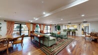 Located at 725 S Bixel St in downtown Los Angeles, this single-floor common space offers a thoughtfully designed layout that balances functionality with classic elegance. Rich hardwood flooring flows throughout the main areas, anchoring the welcoming ambiance from entry to lounge.

The entrance foyer greets visitors with ornate crown molding, lush potted greenery, a large wall-mounted flat-screen TV atop an intricately carved wooden credenza, and glass double doors that allow natural light to filter in from the outdoors.

The expansive lounge area features recessed lighting, a tray ceiling with decorative crown molding, and large windows dressed in warm, patterned curtains that frame views of the exterior courtyard. Comfortable seating arrangements centered around a glass coffee table create an inviting space for socializing or relaxing.

The kitchen is fully equipped with stainless steel appliances — including a gas range, built-in microwave, dishwasher, and refrigerator — complemented by light maple cabinetry, dark granite countertops, and tile flooring. A bar-height counter provides additional seating.

The bathroom is finished with decorative tilework, a large framed mirror, warm wood vanity cabinetry, and accessible fixtures, reflecting a clean and practical design. Overall, this space exudes a warm, classic character suited for community use.
