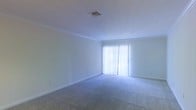 This single-floor residential space offers a clean, well-maintained interior defined by simplicity and functionality. The living area serves as the centerpiece of the layout, featuring plush wall-to-wall carpeting in a neutral gray tone that adds warmth and comfort underfoot. Crisp white walls are accented by crown molding, lending a polished, finished look to the space. A sliding glass door fitted with vertical blinds serves as a prominent architectural feature, flooding the room with abundant natural light while providing direct access to the outdoors. The ceiling is smooth and bright, complemented by a centrally positioned HVAC vent that ensures efficient climate control throughout the space. The open, unobstructed floor plan allows for flexible furniture arrangement to suit a variety of living configurations. White baseboards frame the perimeter of the room, reinforcing the clean aesthetic that runs consistently through the interior. The overall ambiance is airy, neutral, and move-in ready, making it a versatile space well-suited for a range of personal styles and furnishing preferences. The abundance of natural light, combined with the light color palette, creates an inviting and refreshing atmosphere throughout.