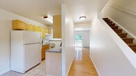 Located at 210 Vineyard Ave in Highland, NY, this two-story home offers a well-organized layout across two floors. The main level houses the kitchen and living room, connected in an open-concept arrangement. The kitchen features light wood cabinetry, a peninsula with a granite-style countertop that provides a breakfast bar overhang, tile flooring, and standard appliances including a full-size refrigerator and electric range. A pendant light adds a functional accent above the kitchen area. The living room extends from the kitchen with warm hardwood-style flooring and is brightened by a sliding glass door that opens onto a wooden deck overlooking lush greenery, allowing for excellent natural light and outdoor access. A hallway and staircase connect the main floor to the upper level. Upstairs, two bedrooms offer comfortable accommodations, each with soft carpeting, baseboard heating, and natural light from windows. The primary bedroom includes a closet and ample floor space. The full bathroom is finished with warm earth-tone tile on both the floor and tub surround, complemented by a white vanity with cabinet storage and a mirrored medicine cabinet, providing a clean and functional design throughout.