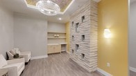 This single-floor space showcases a refined blend of modern design elements and warm, inviting aesthetics. The centerpiece of the main area is a dramatic floor-to-ceiling stacked stone accent wall, finished in light cream and beige tones, featuring a series of recessed display niches at varying levels — ideal for showcasing décor or functional storage. Warm golden-yellow accent walls contrast beautifully with the soft neutral grey of the primary walls, creating a balanced and welcoming atmosphere. Underfoot, wide-plank laminate flooring in a light wood tone runs throughout, adding warmth and continuity to the space. The ceiling draws immediate attention with a coffered tray detail adorned with an ornate patterned inset, from which a stunning crystal chandelier hangs, casting brilliant ambient light across the room. A built-in workstation along one wall features light maple cabinetry and open shelving, offering both practicality and a clean, streamlined look. Recessed lighting complements the chandelier, ensuring the space is well-lit at all times. Together, these elements create an environment that is both functionally equipped and visually sophisticated, suitable for professional or hospitality use.