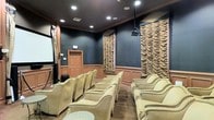 This single-floor private screening room is thoughtfully designed to deliver a premium cinematic experience. The room is appointed with rows of generously sized, cream-colored upholstered armchairs accented with nailhead trim, offering comfortable, lounge-style seating for a sizeable audience. The walls are finished in a deep navy-blue above classic wainscoting, lending the space a sophisticated, theater-inspired aesthetic. Dramatic textured acoustic panels — featuring layered, wave-like burlap or fabric forms — are mounted along the side walls, serving both as sound-dampening elements and striking decorative features that enhance the room's refined character. Recessed ceiling lighting provides subtle, controlled illumination, while a ceiling-mounted projector points toward a large projection screen at the front of the room. Crown molding ties the design together, adding an elegant finishing touch. A solid wood entry door with a transom provides discreet access to the space. The overall ambiance is intimate yet polished, combining the comfort of a luxury lounge with the focused functionality of a dedicated home or private theater.