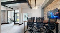 Located at 2201 NW Federal Hwy N in Stuart, Florida, this single-floor commercial office space offers a thoughtfully designed environment that blends modern aesthetics with functional workspaces. The layout encompasses an open lounge area, a collaborative conference room, private offices, and a utility nook, all unified by a cohesive industrial-chic design language.

The main lounge area features large-format light tile flooring, teal-accented structural columns, and an abundance of natural light streaming through floor-to-ceiling windows that frame views of the surrounding greenery. Comfortable seating arrangements, including leather sofas and accent chairs, create an inviting atmosphere for informal gatherings.

A dedicated conference room anchors one end of the space, furnished with a rich walnut-toned table and ergonomic mesh chairs. Exposed black ductwork runs along the ceiling alongside a striking cluster chandelier, lending an urban, loft-inspired character. A sliding barn-style glass door and a wall-mounted display screen complete the room.

A secondary private conference room features floor-to-ceiling glass partition walls with black framing, a clean wood-topped table, and large windows overlooking lush outdoor landscaping, filling the room with natural light.

A textured brick accent wall adds warmth throughout, while a carpeted waiting alcove with upholstered lounge chairs and a wooden side table provides a welcoming reception area. An adjacent workroom with a decorative wallpapered accent wall houses essential office equipment.