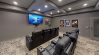 This private theater is thoughtfully designed to deliver an immersive cinematic experience from the comfort of home. The room is anchored by a large wall-mounted flat-screen display positioned at the front, providing an unobstructed sightline from every seat in the house. Multiple rows of plush black leather power recliner seats — each equipped with built-in cup holders and armrests — are arranged in a tiered layout, ensuring optimal viewing angles throughout the space.

The ceiling is a standout architectural feature, showcasing an elegant coffered design with recessed lighting that casts a warm, ambient glow across the room. The neutral gray walls are tastefully adorned with framed classic and contemporary movie posters, reinforcing the theatrical atmosphere. A decorative sunburst wall accent and a built-in back counter area add refined finishing touches to the rear of the room.

Patterned carpeting in soft, muted tones runs throughout, contributing to both the aesthetic warmth and acoustic quality of the space. Roller shades on the windows allow for effective light control when the screen is in use. A dedicated entry door and a small adjacent utility area complete the layout, making this theater a fully self-contained entertainment destination designed for serious film enthusiasts.