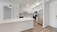 This single-floor residence offers a clean, contemporary design with a bright and open layout. The heart of the home is a well-appointed kitchen featuring crisp white cabinetry extending to the ceiling, a subway tile backsplash, a large central island with a white countertop, pendant lighting, and a full suite of stainless steel appliances including a refrigerator, electric range, and microwave. The kitchen flows seamlessly into a spacious living area finished with light-toned wood-look flooring, creating a cohesive and airy atmosphere throughout the shared space. A secondary living or dining area is also finished with the same wood-look flooring and features direct access to an outdoor space through a glass door, allowing for natural light to fill the room. The bedrooms are carpeted for comfort and feature large windows with blinds that invite generous natural light while providing privacy. Two full bathrooms are finished with wood-look flooring, white vanity cabinetry, white countertops, large mirrors, and tub-shower combinations. One bathroom includes accessibility grab bars. The overall aesthetic throughout is modern and neutral, with white walls and clean lines creating a fresh, move-in-ready environment.