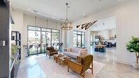 This contemporary clubhouse in Denham Springs, Louisiana, delivers a striking first impression through its grand entry foyer, where soaring vaulted ceilings with exposed wood beam accents and large-format black-framed glass doors set a bold architectural tone. Polished concrete floors flow seamlessly throughout, tying together the open-concept layout with a clean, modern aesthetic.

The expansive main gathering area is bathed in natural light through floor-to-ceiling windows that frame views of the surrounding community. Warm pendant chandeliers and statement lighting fixtures complement the natural wood and woven textures found in the carefully curated furnishings, including rattan accent chairs, live-edge driftwood coffee tables, and plush seating arrangements. A large wall-mounted television and built-in display shelving anchor one end of the lounge, while a high-top bar area runs along the windowed perimeter.

A fully equipped kitchen features gray shaker cabinetry, white quartz countertops, a farmhouse sink, subway tile backsplash, floating wood shelves, and premium stainless appliances. Adjacent dining and lounge zones provide flexible seating options for gatherings of all sizes.

Glass-paneled sliding doors lead to private rooms suited for meetings or events. Curated artwork, including a dramatic landscape triptych, adds a refined, gallery-like quality to the overall ambiance of this well-appointed community amenity space.