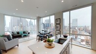 This sophisticated single-floor urban residence showcases a seamlessly integrated open-concept layout that combines the kitchen, dining, and living areas into one cohesive and elegant space. Floor-to-ceiling glass windows wrap around the unit on multiple sides, flooding the interior with abundant natural light and framing breathtaking panoramic views of the surrounding city skyline — a true centerpiece of the design.

The modern kitchen features a generous island with a crisp white countertop and an undermount sink, complemented by sleek cabinetry with stainless steel hardware. The island serves as both a functional workspace and a natural gathering point for the open living area. A compact dining area with a dark-toned table and matching stools sits adjacent to the kitchen, perfectly positioned to take advantage of the dramatic cityscape views.

The living area extends naturally from the dining space, offering a comfortable seating arrangement set atop a soft area rug. Recessed lighting throughout the ceiling complements the natural light, ensuring the space remains bright and inviting at any time of day. Exposed concrete columns add an industrial-chic contrast to the otherwise clean, neutral palette of whites and warm beiges. The overall ambiance is refined, airy, and decidedly metropolitan.