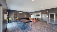 Located at 2800 New Brunswick Ave in Piscataway, NJ, this single-floor community lounge is a well-appointed recreational and social gathering space. The interior is defined by a contemporary design aesthetic, featuring dark charcoal-painted walls, wood-look plank flooring, and recessed lighting throughout that creates a sleek, modern ambiance.

The open layout is organized into distinct activity zones. The game area serves as the centerpiece of the space, anchored by a full-size pool table and a shuffleboard table, complemented by wall-mounted motivational signage that reinforces the room's energetic character. A high-top bar table with seating is positioned nearby, ideal for spectators and socializing.

A comfortable lounge area occupies one end of the room, furnished with a large sectional sofa adorned with colorful accent pillows, a coffee table, and a wall-mounted flat-screen television. Multiple large windows in this area flood the seating zone with natural light and offer views of the outdoor surroundings, including what appears to be a pool area.

On the opposite end, a wet bar area with cabinetry and a sink provides a convenient refreshment station, accompanied by an additional wall-mounted television. The space is equipped with a wall-mounted mini-split HVAC unit, ensuring year-round climate comfort. Multiple entry points, including glass-paneled doors, provide easy access to the outdoors.