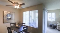 This single-floor residence offers a functional and comfortable living environment with a clean, practical design. The dining area is a standout feature, bathed in natural light streaming through large windows dressed with vertical blinds. A ceiling fan with an integrated light fixture is centrally positioned overhead, providing both airflow and ambient lighting. The dining space comfortably accommodates a dark-toned wooden table and matching chairs, creating a welcoming setting for meals and gatherings. Neutral-toned walls complement the warm flooring, contributing to a cohesive and understated aesthetic throughout. A sliding glass door adjacent to the main window provides direct access to an outdoor patio area, where additional seating is visible, extending the living space outdoors. The textured ceiling adds a subtle architectural detail consistent throughout the space. Overall, the residence balances practicality with comfort, featuring ample natural light, easy indoor-outdoor flow, and a neutral palette that offers a versatile backdrop for a variety of interior styles.