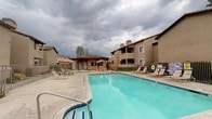 This single-story community amenity space offers residents access to both an indoor fitness center and a well-appointed outdoor pool area. The fitness center is a bright, open room finished with neutral-toned carpet, light gray walls, and recessed lighting throughout. It is equipped with a treadmill, stationary recumbent bikes, and an elliptical trainer, providing a solid range of cardio equipment for residents. Large sliding windows line the walls, allowing natural light to fill the space and offering direct views of the outdoor pool area, creating a connected and airy atmosphere. A dedicated entry with tile flooring and a small service counter area is also present within the fitness room. The outdoor area showcases a generously sized swimming pool with clear blue water, accompanied by an adjacent spa. A wide pool deck surrounds the water features and is furnished with lounge chairs for relaxing. A covered gazebo structure with a pitched tile roof provides shaded seating nearby. The community buildings visible from the pool area display a Mediterranean-influenced architectural style, featuring tan stucco exteriors, terra cotta tile rooflines, and wrought iron fencing enclosing the pool area for safety and privacy.