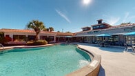 Located along U.S. 98 in Pensacola, Florida, this single-story outdoor amenity complex offers a resort-style experience designed for relaxation and recreation. The centerpiece is a generously sized freeform swimming pool with a smooth curved edge, surrounded by a broad concrete sun deck lined with lounge chairs and blue market umbrellas that provide ample shade throughout the day. The pool area is framed by a Mediterranean-inspired clubhouse building featuring a distinctive red tile roof, large floor-to-ceiling windows, and French doors that open directly onto the poolside patio — allowing natural light to flood both indoor and outdoor spaces seamlessly. Manicured landscaping, including palm trees and trimmed shrubs set against mulched garden beds, adds a tropical character to the environment. A separate, expansive pool with an extended sundeck and additional rows of lounge seating provides further space for recreation, backed by a natural tree line that offers privacy and greenery. A covered gazebo pavilion with ceiling fans, bar-height seating, and column detailing serves as a social gathering point within the complex. The grounds also include a playground area and open grassy spaces, completing a well-rounded outdoor amenity hub suited for community use in the Florida sunshine.
