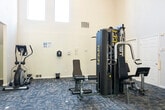 This fitness center is a thoughtfully designed, single-floor facility offering a spacious and welcoming environment for a full range of workout routines. The main workout area is immediately distinguished by its dramatically vaulted ceilings and tall, narrow windows that flood the space with natural light, creating an open and airy atmosphere. Patterned blue-grey carpeting covers the floor throughout, providing both comfort and durability underfoot.

The facility is divided into two connected zones. The primary area houses a multi-station cable weight machine, an elliptical trainer, a stationary upright bike, and a weight bench, offering users a variety of strength and cross-training options. A set of glass double doors along one wall opens to an outdoor area, further enhancing the sense of openness and ventilation.

A secondary alcove-style area, accessible through an arched opening, features a treadmill, an additional elliptical trainer, a stationary recumbent bike, and a vertical knee raise/dip station. This lower-ceilinged section is lit by flush-mount ceiling fixtures and benefits from its own window, maintaining a bright and functional atmosphere.

Together, the two zones provide a well-rounded fitness experience, catering to both cardio and strength training needs in a clean, organized, and light-filled setting.