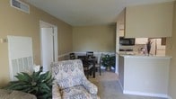 This single-floor space offers a warm and functional layout, highlighted by an open-concept design that seamlessly connects the living, dining, and kitchen areas. The living area is finished with plush wall-to-wall carpeting that adds a soft, comfortable feel underfoot, while neutral warm beige walls create a calming and versatile backdrop throughout. A decorative chair rail runs along the lower portion of the walls, adding a subtle architectural detail that lends a classic touch to the space. The living area provides enough room for comfortable seating arrangements, as seen with the patterned accent armchair, and is complemented by the presence of indoor greenery that adds a natural, welcoming element. The dining area accommodates a small table with chairs, making it suitable for intimate meals. Adjacent to the dining space, a compact kitchen is visible, featuring essential appliances including an over-the-range microwave and stovetop. A light-colored countertop bar divides the kitchen from the main living space, offering both functionality and an open sightline. Doorways visible from the main area suggest additional private spaces beyond. Overall, the layout is efficient and inviting.